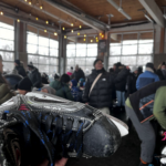 A hockey skate being held up in front of a packed room of people with twinkly lights and lots of windows.